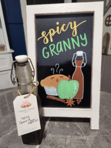 spicy granny apple simple syrup in glass bottle in front of sign with syrup name, apple pie, a green apple, and the syrup bottle drawn in chalk